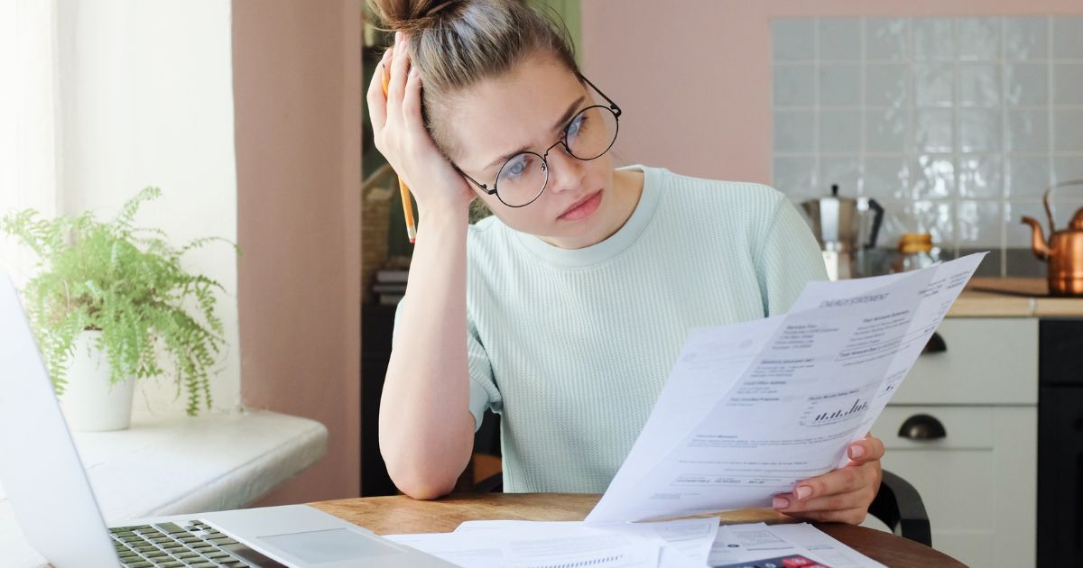 Mujer con piel blanca y pelo rubio con espejuelos confundida leyendo un papel de análisis financiero